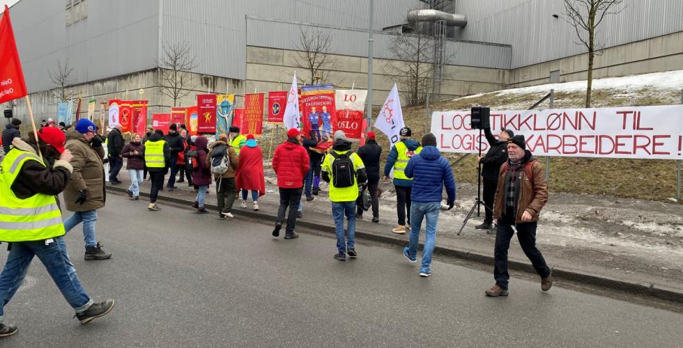 Mennesker g&aring;r over veien, skilt og bannere i bakgrunnen
