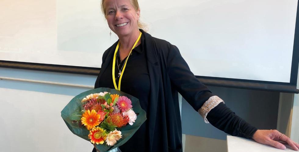 Her er Tonje, ei dame som st&aring;r foran en prosjekter med en blomsterbukett i h&aring;nda. Hun har p&aring; seg svarte kl&aelig;r, og har h&aring;ret i hestehale. 