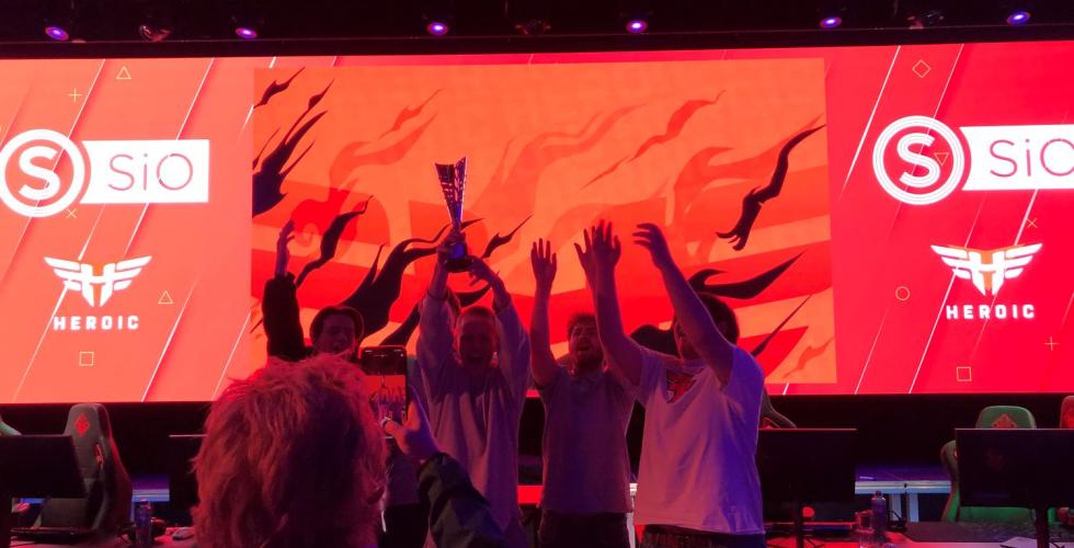 Fem jublende personer st&aring;r foran p&aring; en scene og l&oslash;fter en pokal. 